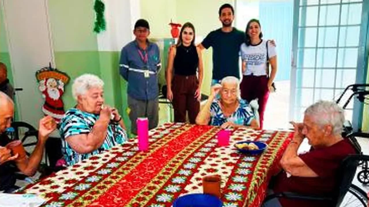 Idosos à mesa de café da manhã com colaboradores da Santin em pé ao fundo, na Casa do Idoso de Américo Brasiliense (SP)
