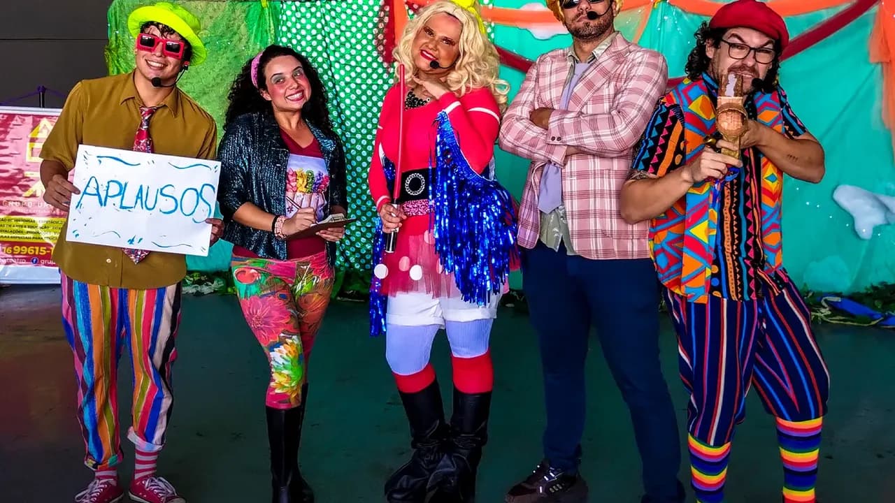 Atores fantasiados em apresentação de teatro, um deles segurando cartaz com a palavra Aplausos