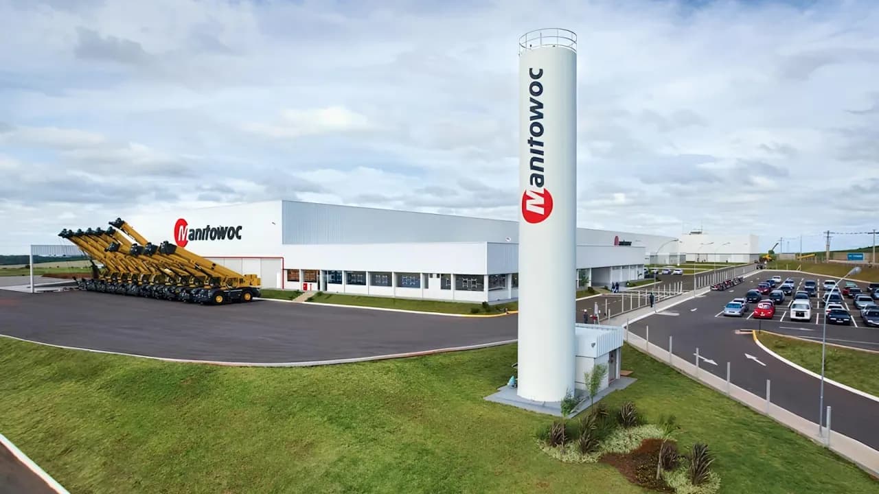 Fábrica da Manitowoc com torre com o logotipo da empresa e guindastes amarelos estacionados em frente ao galpão