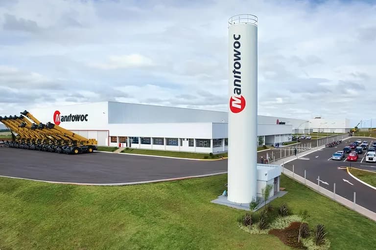 Fábrica da Manitowoc com torre com o logotipo da empresa e guindastes amarelos estacionados em frente ao galpão
