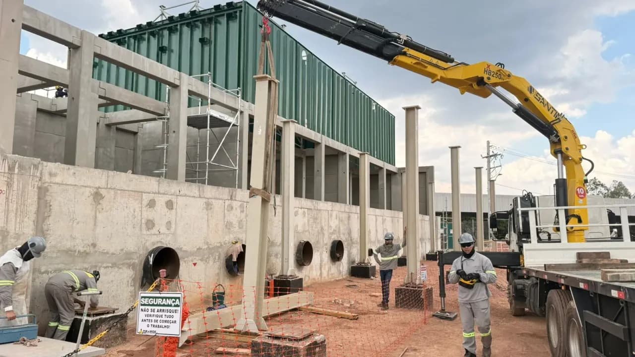 Caminhão com guindaste articulado amarelo descarregando peças no canteiro da URE Barueri, com equipe de montagem